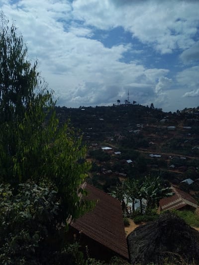 The image displays a panoramic view of a densely populated, hilly urban environment identified as the city of Kilambo, Democratic Republic of the Congo, under a partially cloudy sky. In the foreground, a tall, slender tree with light green, elongated leaves occupies the left side, partially obscuring the lower view. To the right, a dark brown corrugated metal roof of a building is visible, sloping downwards, adjacent to a dark, dome-shaped mound of vegetation, possibly thatch or piled organic material. The midground and background reveal numerous residential structures with varied roofing materials, primarily corrugated metal, spread across undulating terrain. Patches of reddish-brown earth, indicative of exposed soil or unpaved paths, are visible between structures and interspersed with green trees and vegetation. On a prominent distant hilltop, a tall communication tower, likely a cellular or radio mast, stands out against the sky, accompanied by smaller structures. The sky above features a mix of white and grey cumulus clouds with visible patches of blue. No individuals or specific actions are discernible within the frame.
