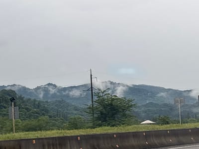 The image depicts a landscape with rolling, mist-shrouded mountains in the background, indicative of the environment in the region of ნიგოითის თემი, Georgia. In the foreground, a concrete barrier separates a grassy area from what appears to be a roadway. Several tall utility poles with power lines traverse the scene, one of which is prominently positioned in the middle ground. There are also two road signs, one on the left and another on the right, partially obscured by vegetation. A small, white, conical-roofed structure is visible amidst the trees on the right side of the frame. The overall atmosphere is overcast, with a soft, diffused light.