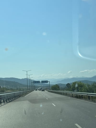 A clear, bright day captures a highway scene with mountains in the distance. The road is paved and appears to be a multi-lane highway, with guardrails lining both sides. Several cars are visible on the road, driving in the same direction as the viewer. Overhead, blue signs with white lettering and arrows indicate directions to various locations, including "Novachene" and "Sofia." The sky is a light blue with a few scattered white clouds. Green trees and hills are visible on either side of the highway, leading up to the more distant, layered mountain range. The overall impression is one of travel and open road on a sunny day.