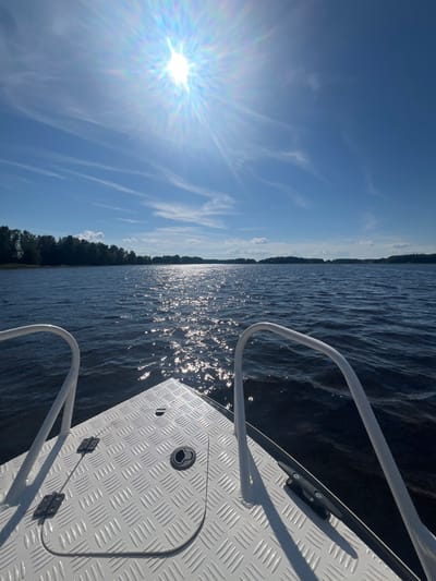 This is a slightly tilted, eye-level, outdoor, medium shot of a sunny day on a body of water, likely a lake, in Сюктерка, Russia. The sun is bright and low in the sky, casting a strong glare on the water's surface and creating a lens flare in the sky. The sky is a clear blue with wispy white clouds. The water is dark and choppy, with numerous bright reflections of the sun dancing on its surface.
In the foreground, the edge of a white boat is visible, featuring a diamond-plate metal deck and a white metal railing. A black cleat is mounted on the deck, and a small, hinged compartment with a circular latch is also visible. The railing curves away from the viewer, suggesting movement or the shape of the boat's bow.
In the distance, a dense line of dark green trees borders the water, indicating a forested shoreline. The overall scene evokes a sense of peacefulness and the enjoyment of a warm, sunny day on the water. The time of day appears to be late morning or early afternoon, given the sun's position and the brightness of the light. There is no visible text in the image.