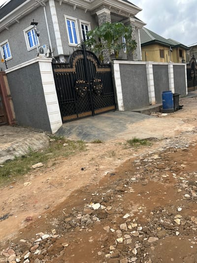 This image depicts a residential street scene in Ashipa, Nigeria, under an overcast sky. In the foreground, the ground consists of reddish-brown dirt and gravel, intermingled with scattered white and grey fragments of debris, and sparse patches of green grass. A paved concrete surface serves as a driveway leading to a dark grey perimeter wall, featuring white vertical decorative pillars. Centrally positioned in the midground is a large, ornate black metal double gate with gold-colored scrollwork, providing access to the property.

Behind the gate and wall, a two-story residential building dominates the left background. It exhibits a grey textured facade with white trim, multiple windows fitted with blue-tinted glass, and a dark grey roof. A white outdoor air conditioning unit and a black and white surveillance camera are mounted on the exterior wall of this building, to the left of the gate. To the right of the primary property, a section of another residential structure is partially visible, characterized by a yellowish-tan facade and a dark green roof, with its perimeter wall topped by electric fencing. A blue plastic barrel, faintly marked "OIL" on its side, and a black waste bin are situated on the paved area near the rightmost section of the perimeter wall. No individuals are present in the scene.