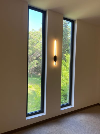 The image depicts an interior view of a room with two tall, narrow windows. A vertical, modern wall sconce with a warm light is mounted on the wall between the windows. The windows offer a view of lush green trees and a grassy area, suggesting an outdoor setting. The interior features beige walls and a textured carpeted floor. The scene is well-lit, with natural light coming from the windows and artificial light from the sconce. The overall impression is one of a contemporary and serene space. The location is identified as Pitsunda, Abkhazia.