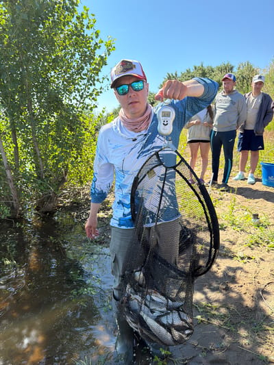 In Tarasovo, Russia, a man in a blue long-sleeved shirt, khaki shorts, and a red and white baseball cap stands by a body of water, holding a fishing net filled with fish. He is also holding a digital scale that reads "6.4". Behind him, three other individuals are visible, along with trees and a bright blue sky. The ground is dirt and grass.