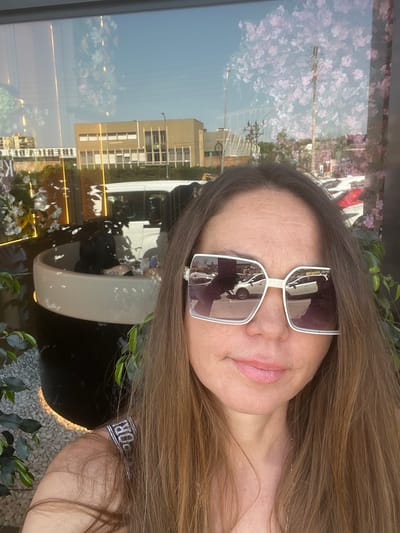 A selfie of a woman with long brown hair wearing white-framed sunglasses. The reflection in the sunglasses shows a street scene with parked cars. The background behind the woman is a glass window reflecting buildings and flowers. Based on the context, the location is Torreforta, Spain. There is a plant and a black object in the foreground. The woman is wearing a garment with a white band visible.