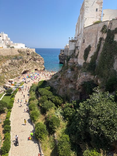 The media file displays a vibrant scene from Polignano a Mare, Italy, on a clear, sunny day. The image captures a bustling beach and a nearby pathway lined with lush greenery.
The primary subjects are the numerous people enjoying the beach and the promenade. On the beach, individuals are sunbathing, relaxing under colorful umbrellas, and wading in the clear blue Mediterranean Sea. The beach itself is a mix of sand and rocky outcrops, with several caves carved into the cliff face. Above the beach, a paved pathway winds along the coastline, populated by people strolling, exercising, and observing the scenic view. Some individuals are pushing strollers or carrying bags, suggesting a leisurely outing.
Notable details include the traditional white-washed buildings perched on the cliffs overlooking the sea, a common architectural style in the region. The contrast between the rugged, natural cliff formations, the man-made structures, and the vibrant blue of the water creates a picturesque setting. The time of day appears to be midday or early afternoon, given the bright sunlight and the shadows cast on the ground. The overall mood is one of relaxation, recreation, and enjoyment of a beautiful coastal environment. There is no visible text in the media file.
