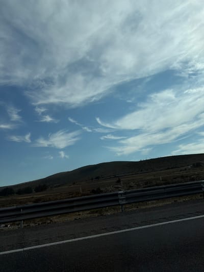 A road runs vertically along the right edge. Adjacent to it, a series of pipes follow the same trajectory. A small hill lines the pipes and road. The majority of the image is filled with the sky, populated by thin, white clouds scattered across a blue expanse. The orientation of the image is such that the sky appears on the left, while the land features are compressed to the right.