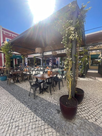 The image presents an outdoor dining patio located within the Freeport Fashion Outlet in Alcochete, Portugal. The scene is illuminated by bright sunlight from a clear blue sky, causing significant lens flare in the upper-center.
A large, dark-colored metal pergola with a slatted wooden roof extends across the foreground and midground, providing shade for an array of empty dining tables. Vines with white flowers are extensively trained around the pergola's dark metal support poles, which emerge from large, dark brown, rounded planter pots filled with soil. One light blue planter pot is visible on the far left.
The patio features numerous outdoor dining setups: rectangular and square tables with dark wooden or composite tops and black metal pedestal bases are surrounded by black metal chairs, many exhibiting an industrial or distressed finish. Some tables hold small orange cylindrical objects.
The ground is paved with traditional Portuguese cobblestones (calçada portuguesa), forming a diamond pattern primarily in light grey, with a distinct longitudinal strip of darker grey cobblestones running through the center of the seating area. Distinct shadows are cast on the cobblestones due to the high sun angle.
In the background, multi-story buildings flank the patio. On the far left, a building with a red and white facade is visible, beneath which a storefront with a green awning is situated. Further back, indistinct human figures are sparsely distributed, walking or standing within the larger complex of shops and eateries.