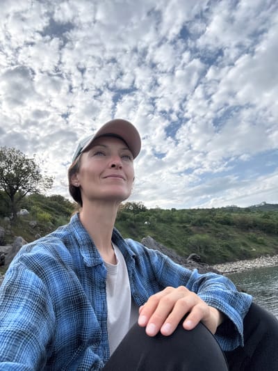 A woman is sitting outdoors on a cloudy day. She is wearing a blue plaid shirt over a white t-shirt and a brown cap. The background consists of a sky filled with clouds and a green, hilly landscape. The woman appears to be on a rocky surface. It is not possible to determine if the location is Bondarenkovo, Ukraine from this image.