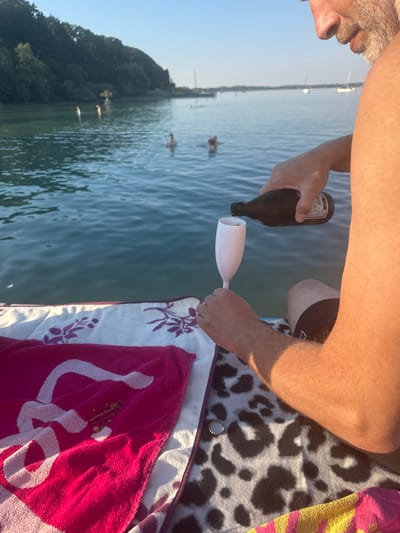 The image shows a person pouring a beverage from a dark brown bottle into a white champagne flute. The scene is set outdoors, on the water, likely a lake given the calm surface and the presence of trees along the shoreline in the background. Sunlight illuminates the scene, suggesting it is daytime, possibly late afternoon due to the warm tones. The person's arm and hand are visible, holding both the bottle and the glass, indicating they are actively engaged in pouring.

In the background, the lake stretches out with a few boats and some people swimming or wading in the water. The shoreline is lined with lush green trees, hinting at a natural or park-like environment. On the right side of the image, parts of towels are visible. One towel has a leopard print pattern in black and white, and another is a bright pink towel with a white floral or paisley-like design. A bottle cap lies on the leopard-print towel.

The overall atmosphere appears relaxed and recreational, consistent with a day spent by the water. The focus on pouring a drink into a champagne flute suggests a celebratory or leisurely occasion. There is no visible text in the image. The location context of Gstadt am Chiemsee, Germany, aligns with the visual cues of a lake setting with a tree-lined shore.
