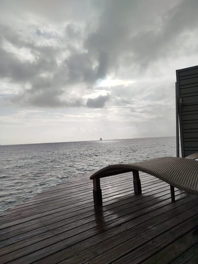 This image captures a serene view of the ocean from a wooden deck. In the foreground, a woven lounge chair is positioned on the wet wooden planks of the deck, with a portion of a dark, slatted structure visible to the right. The ocean stretches out to the horizon under a sky filled with heavy, gray clouds. A small ship is visible in the distance on the water. The scene appears to be in Gulhi Falhu, Maldives, suggesting a resort or private residence setting. The lighting is subdued due to the overcast conditions.