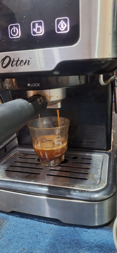 An espresso is being brewed into a glass demitasse cup by an automatic coffee machine. The machine has a black body with a silver metallic finish on the top and front. Above the brewing mechanism, there are three illuminated icons on a black panel. The left icon depicts a power symbol, the middle icon shows a hand pressing a button, and the right icon shows a steaming cup. The brand name "Otten" is visible in white script on the front panel, just below the icons. The coffee machine has a drip tray with horizontal slats, beneath which is a blue cloth. The espresso is dark brown with a thin layer of crema on top, and it is halfway full in the glass. The image appears to be taken indoors, likely in a kitchen or cafe setting, possibly in the city of Rhengbluek, Indonesia, though this cannot be definitively confirmed from the image itself.