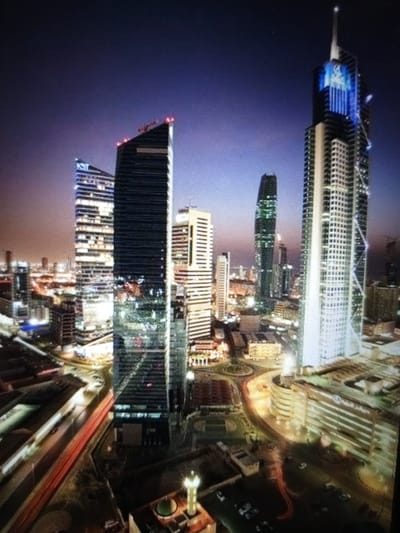 The image depicts a nighttime aerial view of a modern cityscape, characterized by numerous illuminated skyscrapers. The dominant structures are tall, sleek buildings with glass facades, many of which are lit up, creating a vibrant and dynamic scene. The sky above is a deep, twilight purple, transitioning to a lighter shade towards the horizon.
In the foreground, a winding road or highway is visible, with streaks of red light indicating the movement of vehicles, suggesting traffic flow at night. Several buildings of varying heights are clustered together, forming a dense urban landscape. Some of the skyscrapers have distinctive architectural features, such as pointed spires or uniquely shaped tops. One particularly tall building on the right features a prominent blue light display near its summit.
The overall impression is one of a bustling metropolis at night, with a focus on modern architecture and urban infrastructure. The lighting from the buildings and vehicles creates a sense of activity and energy. The image appears to be a photograph, possibly taken from a high vantage point like a helicopter or another tall building.