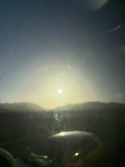 The image displays a sunlit sky with hazy mountain silhouettes in the background. The sun is a bright, white orb, slightly off-center to the right. There is a notable glare across the entire image, indicative of a lens flare or reflection, with streaks of light and dust visible. In the foreground, a dark, out-of-focus object, possibly the dashboard or interior of a vehicle, occupies the lower portion of the frame. The overall impression is of a landscape viewed through a dirty window or with camera artifacts. The location is identified as Puerto del Rosario, Spain.