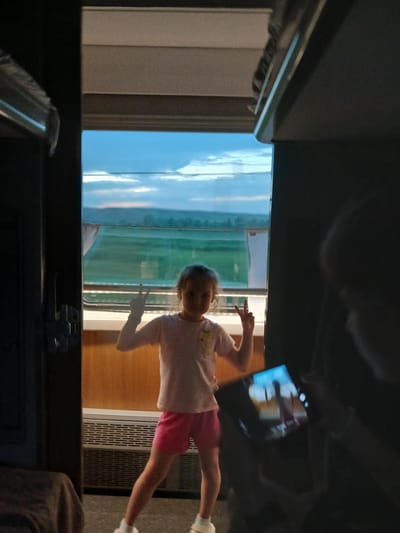 A young girl with blonde hair stands in the doorway of a train car, looking at the camera and holding up two fingers in a peace sign. She wears a light pink t-shirt and pink shorts. Outside the train car window, a green landscape is visible under a cloudy blue sky. To the right, a person is holding a tablet computer, displaying an image of the same girl. The interior of the train car is dimly lit, with wooden paneling and a vent visible on the wall. The overall setting appears to be a train traveling through the countryside near Novo-Yakupovo, Russia.