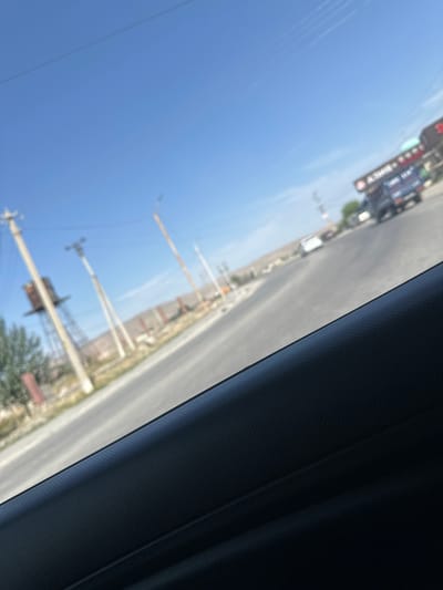 The image depicts a street scene in Ынтымак, Kyrgyzstan, viewed from inside a vehicle. The perspective is tilted, suggesting the photo was taken while driving. The sky is a clear, bright blue, indicating it is daytime, likely mid-morning or afternoon given the sunlight.

The foreground is dominated by the interior of the vehicle, specifically a dark, textured surface that appears to be part of the car's A-pillar or window frame. This dark area takes up a significant portion of the right side of the image.

Outside the vehicle, a road stretches into the distance, bordered by a somewhat sparse landscape. Several utility poles with wires are visible along the roadside, along with some low-lying buildings or structures that are blurred by motion and distance. In the mid-ground, there are signs and what appear to be traffic signals or railroad crossing arms. A few vehicles, including a bus and cars, are also present on the road, contributing to the sense of a populated area. One of the cropped images reveals a sign with text that is partially legible as "2100." The overall impression is one of a typical day in a town or city, with everyday traffic and infrastructure. The scene is static due to the motion blur, but suggests a dynamic environment.