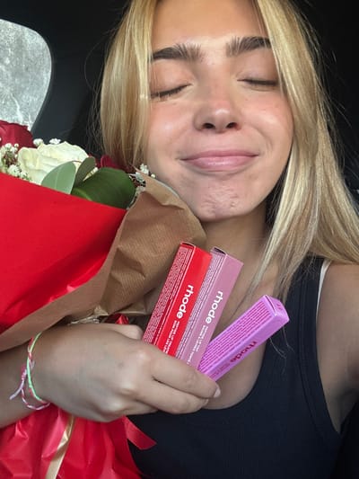 A female individual with blonde hair and a tanned complexion is depicted from the chest up, seated inside a vehicle in Cidade da Maia, Portugal. Her eyes are closed, and her lips are slightly pursed in an expression of contentment. She wears a black ribbed tank top. Her left wrist bears a multi-colored string bracelet. She cradles a bouquet of red and white roses, wrapped in red and brown paper with a red ribbon, in her left arm. In her right hand, held against her chest, are three rectangular boxes of "rhode Peptide Lip Tint" in red, light pink, and purple.