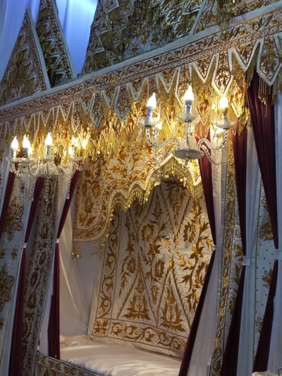 The image shows a close-up view of an elaborately decorated bed, likely part of a traditional Indonesian wedding ceremony, possibly in the city of Cottrueng, Indonesia. The bed is draped with rich fabrics, predominantly white with intricate gold embroidery featuring floral and avian motifs. Burgundy velvet curtains are also part of the draping, adding to the opulent feel. Above the bed, a crystal chandelier with lit bulbs hangs, illuminating the ornate decorations. The overall impression is one of grandeur and cultural significance.