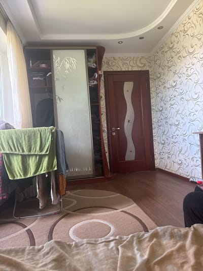 The image depicts an indoor scene, likely a bedroom or living area, in the village of Horzhyly, Ukraine. The room is furnished with a large sliding door wardrobe with frosted glass panels featuring a bamboo design. To the right of the wardrobe is a dark brown wooden door with a decorative wavy glass insert and a silver handle. The walls are covered in patterned wallpaper with a swirling silver design. A wooden floor is visible. In the foreground, a clothes rack is set up, holding several towels and other clothing items, with a light-colored duvet or blanket draped over the edge of the bed in the very front. The ceiling has recessed lighting and decorative molding. Natural light enters from a window on the left, covered by sheer curtains.