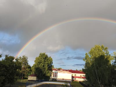 A vibrant double rainbow arches across a cloudy sky over a school or institutional building in Shumerlya, Russia. The primary rainbow is clearly visible, with its distinct spectrum of colors, while a fainter secondary rainbow is discernible above it. The sky is a mixture of dark gray storm clouds and patches of bright blue, suggesting the recent passing of rain. Several small black birds are scattered across the sky, some appearing to be in flight.
In the foreground, lush green trees frame the scene, with a large birch tree prominently featured on the right, its leaves illuminated by sunlight. Below the trees, a white and red building with a red roof is visible. The building appears to be a multi-story structure with numerous windows. In front of the building, there's a sports area, possibly an outdoor ice rink with blue and white borders. A black fence separates the sports area from the surrounding grassy area.
The time of day appears to be late afternoon or early evening, given the angle of the sunlight and the dramatic sky. The presence of the rainbow and the mixed cloud cover indicate changeable weather conditions. The overall mood is one of natural beauty and a sense of calm after a storm. No visible text is present in the image.