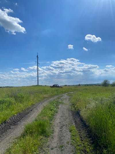 The image shows a dirt road cutting through a grassy field under a bright blue sky with scattered white clouds. A single, tall power line pylon stands in the middle distance, with several power lines stretching across the sky. To the right of the pylon, a rusty red car is parked in the field. The road appears to have tire tracks, suggesting recent use. In the far distance, a treeline is visible, indicating the edge of the field. The overall scene suggests a rural or semi-rural environment.