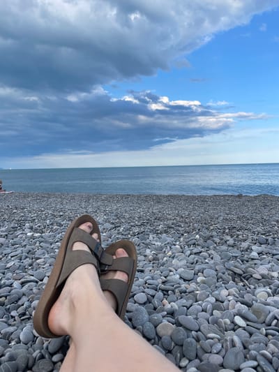 A person's feet, wearing brown Birkenstock sandals, are visible in the foreground, resting on a grey pebble beach. The pebbles are smooth and varied in size and color. Beyond the beach, the calm, blue sea stretches to the horizon, meeting a pale sky. The sky on the left is filled with dramatic, dark grey and white clouds, suggesting approaching weather or a recent storm. On the right, the sky is a clearer, lighter blue. The image is oriented vertically, giving a sense of looking down towards the sea from the shore. The lighting suggests it is either early morning or late afternoon. No text is visible. The setting is a beach in the Livadiysky settlement council, Ukraine. The overall mood is one of quiet contemplation and observation of the natural environment.