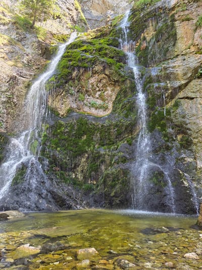 The image depicts a scenic waterfall cascading down a moss-covered rock face. The waterfall is multi-tiered, with water flowing in numerous streams. The surrounding environment appears natural and untouched, with lush green moss covering large sections of the rocks. At the base of the waterfall is a clear pool of water with visible stones at the bottom. Sunlight illuminates the scene, creating highlights on the water and rocks. The context provided indicates this waterfall is located in Landl, Austria. There are no people or animals visible in the image.