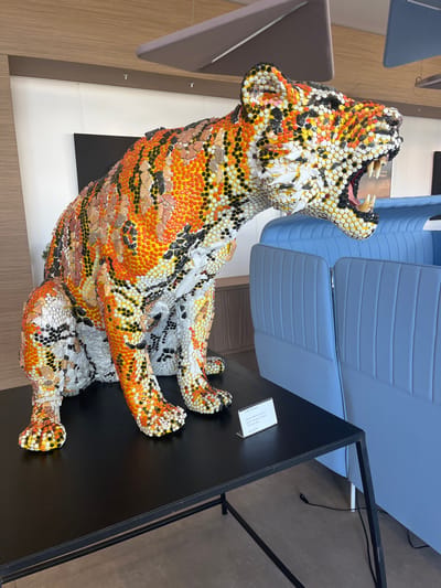 A modern interior in Galatina, Italy, showcases a large, crouching tiger sculpture constructed from a mosaic of numerous small, circular components in orange, yellow, white, and black, forming a striped pattern. The tiger's mouth is open, revealing white teeth. This sculpture rests on a black rectangular display table with thin metal legs. A small white informational plaque, partially visible and titled "CERO D'ARIA," is positioned on the table in front of the sculpture's right paw. To the right and behind the table are light blue, vertically-stitched, upholstered booth-style seating units. The background features light-colored walls, some with horizontal wood paneling, a light brown floor, and a ceiling equipped with suspended grey acoustic panels. An electrical cord is visible on the floor near the blue seating. No individuals are present.