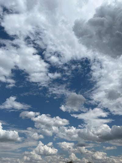 The image shows a sky filled with cumulus clouds against a bright blue sky. The clouds vary in density and shape, with some appearing bright white and others darker and more textured. The lower portion of the image shows a hint of greenery from trees, suggesting an outdoor scene. The overall impression is of a partly cloudy day.