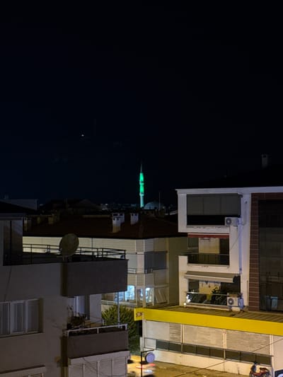 A nighttime, medium-distance, outdoor, vertical shot depicts a dense urban residential and commercial area in Ayvalık, Turkey. The sky is dark with a few faint celestial objects visible. Dominating the skyline is a mosque minaret illuminated in bright green. In the foreground, several multi-story buildings are visible. The leftmost building features a dark, recessed balcony with a satellite dish. Adjacent to it is a building with a red-tiled roof and chimneys. Further to the right is a white building with multiple windows and balconies, one with an outdoor air conditioning unit. The ground floor of this building appears to be a commercial establishment with a yellow awning and shuttered windows. A dark-colored motorbike is parked on the street in the lower right corner. The overall scene is one of urban life at night, with illuminated structures and the distant glow of the mosque.