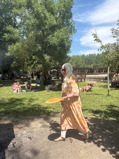 The image depicts a sunny outdoor scene, likely a park or recreational area, with a body of water visible in the background. In the foreground, a woman wearing a hijab and a floral-patterned dress is walking from right to left, carrying a wooden paddle. She is dressed casually in flip-flops. The ground is a mix of dirt and grass, with remnants of a fire pit visible in the lower left corner.

In the middle ground, several people are relaxing on the grass and in chairs. Some are sunbathing, while others are sitting and conversing. Further back, a wooden bridge spans the water, and people can be seen on and around it, some swimming in the water below. The area is shaded by numerous trees, suggesting a warm, pleasant day. The presence of a fire pit and people engaged in leisure activities indicates a picnic or barbecue setting. The natural elements, such as the trees, grass, and water, combined with the relaxed atmosphere, suggest a typical summer day in a park.