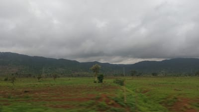 The image depicts a landscape scene, oriented upside down. The upper portion of the image shows a green, rolling field with patches of reddish-brown earth, indicating possibly cultivated land or soil erosion. A line of trees and vegetation runs horizontally across the middle ground, separating the fields from a dark, densely forested mountain range that forms the background. Above the mountains, a thick layer of grey clouds stretches across the sky, suggesting overcast or potentially impending rain.

In the foreground, the sky is a lighter shade of grey, with diffused light indicating either early morning, late afternoon, or a heavily overcast day. The overall mood of the image is subdued and natural, with no discernible human activity or specific objects apart from a lone tree standing prominently in the mid-ground, and a power line tower visible on the left side of the mid-ground. The location context of Gidan Waya, Nigeria, suggests a rural or semi-rural setting, consistent with the natural landscape presented. There is no visible text in the image.