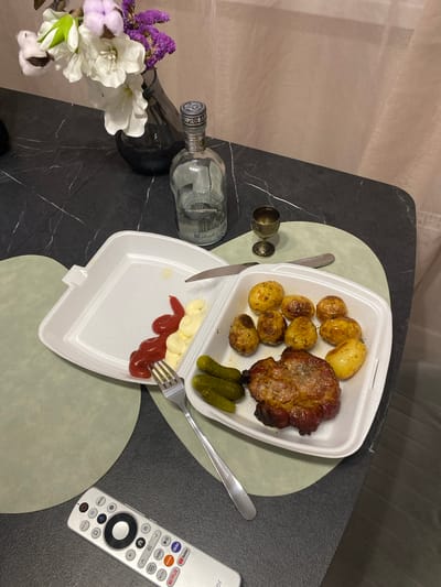 A close-up, overhead shot shows a meal laid out on a dark, marble-patterned table with a light green circular placemat. The meal consists of roasted potatoes and a piece of grilled meat served in a white styrofoam container. Alongside the meat are three dill pickles. A dollop of ketchup and mayonnaise sits on a separate section of the styrofoam container, with a fork resting in the mayonnaise. A knife is placed on the container's edge. To the left of the food, a clear glass bottle of vodka, labeled "РУССКИЙ СТАНДАРТ", lies on its side. Two small, decorative shot glasses are positioned near the bottle. In the lower left corner, a small bouquet of flowers, including purple statice, white amaryllis, and cotton bolls, adds a decorative touch. A silver remote control with various app icons is visible on the right side of the table. The scene is lit by artificial light, suggesting it is indoors, likely in the evening. The setting appears to be a home dining area in Myskhako, Russia.