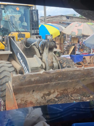 This image depicts a construction site or a street scene in Okha, Nigeria, with a large yellow front-end loader dominating the foreground. The loader's bucket is filled with dirt and debris, and its hydraulic arm is visible. The machine has "SHANTUI" written on its side. A person wearing a hard hat is seated in the driver's cab. In the background, a market area is visible with stalls displaying various goods and covered by umbrellas, one of which is yellow and blue. Buildings with corrugated metal roofs are also present, suggesting a developing urban or semi-urban environment. The overall scene is dusty and appears to be in a busy area with commercial activity alongside the construction equipment.