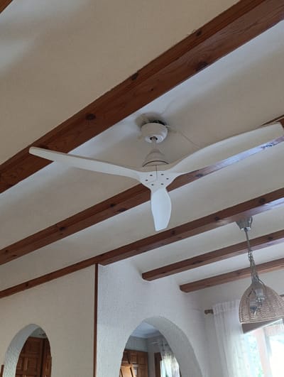 The image shows the interior of a room with a white ceiling supported by exposed wooden beams. A white modern ceiling fan is mounted centrally on the ceiling. To the right of the ceiling fan, a decorative woven lampshade hangs from a chain. The walls appear to be textured and white, with arched openings visible at the bottom of the image, revealing wooden doors. The overall impression is of a well-lit, possibly residential or commercial space with rustic architectural elements. The location is identified as Els Poblets, Spain.