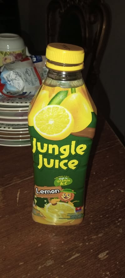 A bottle of Jungle Juice, lemon flavor, stands on a wooden surface. The bottle features a green background with images of lemons. The label highlights that the juice is "High In Vitamin A+C". A cartoon lion wearing a hat appears near the bottom. A yellow cap seals the bottle. Stacked plates and a mug are visible in the background, and a dark chair is in the distance.