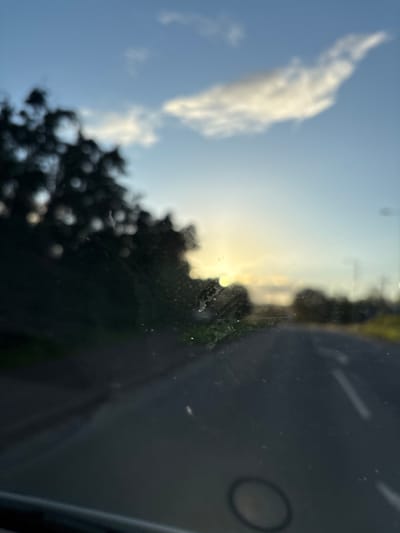 The image shows a view through a car windshield, likely in or near Oswestry, United Kingdom. The sky is visible with a large white cloud and some blue sky. The sun is setting or rising, with bright light shining through trees in the middle ground. The foreground consists of the car's dashboard and windshield, with some visible imperfections on the glass. A road is visible to the right of the frame.