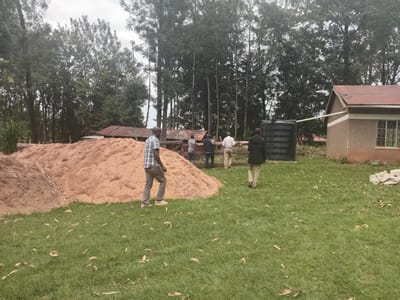 **Location:** Idakho South ward, Kenya.
**Environment:** A grassy field with scattered leaves. Tall trees are visible in the background. A small building with a red roof and an adjacent water tank are present.
**Objects:** A large pile of reddish-brown material (possibly sawdust or sand), a building, a water tank, a fence.
**People:** Five men are visible. One man in a checkered shirt and trousers is walking away from the pile of material. The remaining four men are standing near the fence. One is wearing a dark jacket, another is wearing a light colored shirt.
**Actions:** The men are interacting in some way, presumably talking.