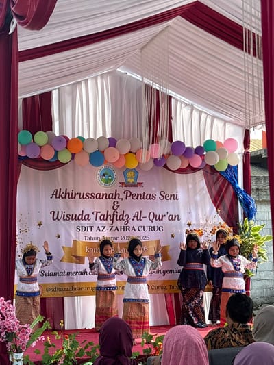 A group of young girls in traditional clothing are performing on a stage in front of a banner that reads "Akhirussanah, Pentas Seni & Wisuda Tahfidz Al-Qur'an SDIT AZ-ZAHRA CURUG Tahun Ajaran 2025-2026". The banner is decorated with balloons and streamers. The girls are wearing colorful outfits with intricate patterns, and some have headpieces. They are all facing the audience with their hands raised. In the foreground, there are flowers and plants. The background shows a tent with red and white fabric. The event appears to be a graduation ceremony or performance for an Islamic school. The location is identified as Sukabakti, Indonesia, based on the banner text which mentions "Curug, Tangerang", a known area within Sukabakti. The audience members are partially visible in the lower part of the image, with one person in a batik shirt in focus.