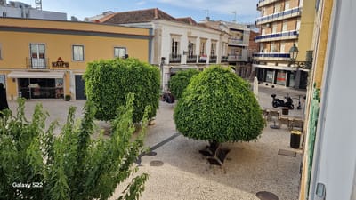 The image shows a public square in Faro, Portugal, paved with cobblestones. In the foreground, there is a lush green plant. Beyond the plant, there are several large, neatly trimmed topiaries. In the background, a yellow building with a restaurant named "KAIFU TABLE BBQ" is visible. Adjacent to it, a white building with balconies and windows is present. Further in the background, a modern building with blue and white striped facade stands tall. Outdoor seating arrangements with chairs and tables are scattered across the square. A black scooter and an electric scooter are parked in the square. A street lamp with ornate metalwork is attached to the building facade. The sky is clear and blue.