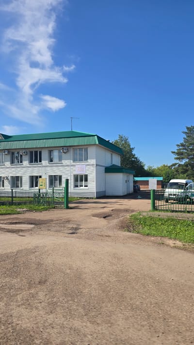 A two-story building with a green, corrugated metal roof stands under a clear blue sky with scattered white clouds. The building's exterior is clad in white horizontal siding. The lower floor has several windows, and the upper floor has more windows, with air conditioning units mounted outside some of them. A sign with the number "70" and some accompanying graphics is visible on the upper facade. A green metal fence with vertical bars encloses a portion of the property in front of the building, with a green metal gate. A dirt road with visible potholes leads to the fenced area. To the right of the building, a white van is parked behind the fence. Trees and other foliage are visible in the background. The overall scene appears to be an industrial or commercial property in the town of Актюбинский, Russia.