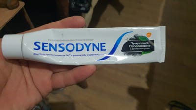 A tube of Sensodyne toothpaste is held in a person's left hand, partially obscuring the label. The toothpaste tube is white with blue lettering and features an image of charcoal and leaves. The text on the tube is in Russian and advertises "№1 по рекомендации стоматологов" (Recommended by dentists #1), "SENSODYNE", and "Природное Отбеливание с древесным углем" (Natural Whitening with charcoal). A small orange GSK logo is visible on the right side of the tube. The background is out of focus and appears to be an indoor setting with a wooden surface and dark shadows. There is no information in the image to suggest the location is Karankul, Uzbekistan.