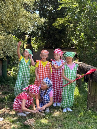 Six young girls, dressed in matching red, white, green, and orange gingham outfits and headscarves, are posed outdoors. The environment appears to be a garden or yard with trees and grass. One girl stands on the left, waving, while another in the center holds a red broom. Two girls are crouched down in the foreground, one examining the ground. The scene is lit by sunlight, indicating it is daytime. The photo is taken in Gderot Regional Council, Israel.