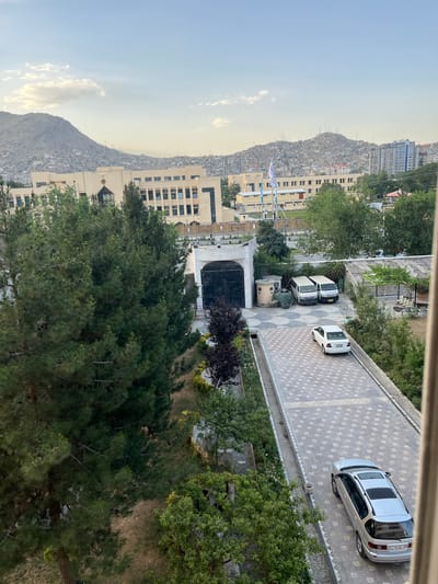 This is an eye-level, outdoor, daytime, long shot of a complex with buildings, trees, and vehicles in Kabul, Afghanistan. The scene is likely the University of Kabul, given the architecture and the flags visible. In the background, the city sprawls across rolling hills, with more distant mountains under a clear sky with scattered clouds. A large, light-colored building with many windows dominates the middle ground, flanked by trees. In the foreground, a paved driveway leads past parked vehicles.

The paved driveway is made of interlocking stone pavers with a diamond pattern. Two white vans are parked near a large, dark gate. A light-colored sedan is parked further down the driveway. To the right of the driveway, there's a covered patio area with seating. The area is bordered by trees and landscaping. There are several people visible in the image, some near the trees on the right side of the frame and others near the vehicles in the driveway. The overall impression is one of a calm, albeit slightly dusty, urban environment.