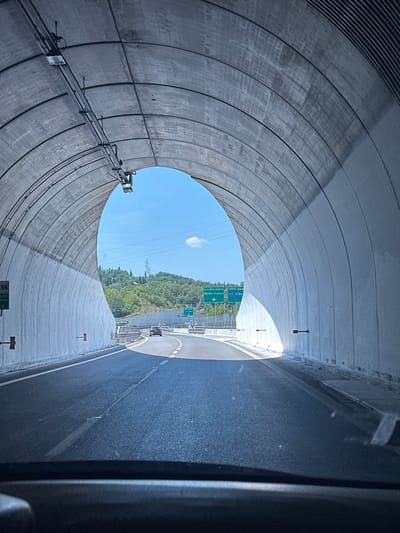 The image shows a view from a vehicle traveling through a tunnel on a sunny day. The tunnel has a curved concrete ceiling with lights and cables running along its length. The exit of the tunnel reveals a clear blue sky with a few clouds and a landscape of green hills covered in trees. There are traffic signs visible, one indicating "BOLOGNA" and another pointing towards "SIENA". A red car is seen driving ahead on the road. The road surface is asphalt with white lane markings. The overall environment appears to be an Italian highway, possibly near Impruneta, Italy, given the directional signs.