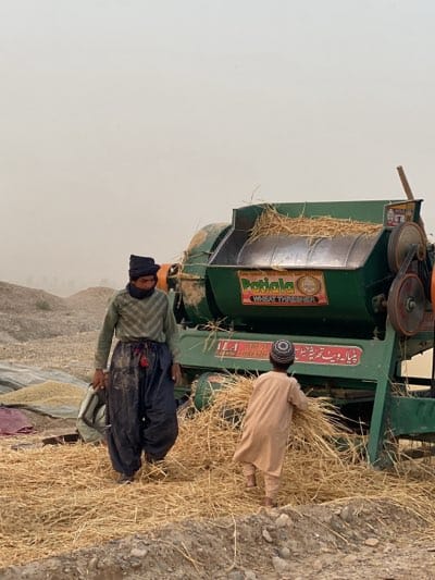 In Alaqadari Daman, Afghanistan, an adult male wearing a black hat, neck covering, and a green and black chevron-patterned shirt paired with dark, loose-fitting pants stands near a green wheat thresher labeled "Patiala Wheat Thresher." The man's pants appear soiled. A child in a tan tunic and patterned cap is in front of the thresher feeding it straw. Loose straw covers the ground around the machine and the two figures. In the background, there is a pile of grain covered with a purple cloth. The sky is overcast, suggesting a dusty or hazy atmosphere.