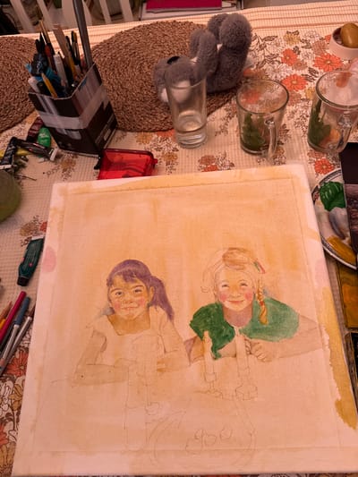 The image depicts an art studio setting on a table. A partially completed watercolor painting of two young girls is the central focus. The girl on the left has dark hair in a ponytail and is wearing a light-colored shirt, with her face partially rendered. The girl on the right has blonde hair with braided pigtails and is wearing a green shirt, her face also partially completed. Both girls have rosy cheeks. The painting is on a canvas, with faint pencil lines visible for the rest of the composition, which appears to include candlesticks and potentially other objects on a table.
Scattered around the painting are various art supplies. To the left, a collection of paint tubes, including a green tube, and colored pencils are visible. In the background, a cardboard container holds an assortment of paintbrushes and pens. Several drinking glasses, some containing what appears to be greenery, are also on the table, along with a red pencil sharpener. A small, gray plush toy is visible behind the glasses. The table is covered with a floral-patterned tablecloth, and a woven placemat is partially visible. The scene appears to be indoors, possibly in a home studio in Kfar Vradim, Israel.