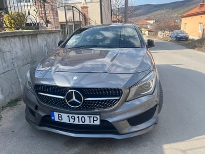 A dark grey Mercedes-Benz CLA-Class sedan is parked head-on on a paved road in the city of Vetren, Bulgaria. The vehicle displays a Bulgarian license plate "B 1910 TP" on its front bumper. A circular Mercedes-Benz emblem is centered on a grille with a diamond-pin design. The front left side of the bumper has a rectangular recess where a cover or sensor is missing. To the immediate left of the car is a grey concrete wall, topped by a black ornate metal fence and gate, behind which a light-colored building is partially visible. The road continues into the background, where an orange-colored house stands on the right, and a blue hatchback is parked further down on an unpaved section. Distant hills or mountains, sparsely covered with trees, form the horizon under a bright blue sky with scattered white clouds. The scene is lit by strong sunlight, casting shadows onto the road surface. No individuals are visible.
