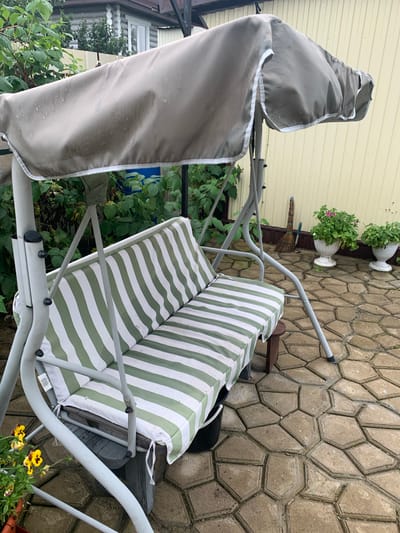 The image shows a garden swing set with a green and white striped cushion. The swing set has a beige canopy that appears to be wet, suggesting it may have recently rained. The swing is on a paved patio made of interlocking hexagonal stones. In the background, there is a light yellow wooden fence, a log house, and some greenery. Two white planters with pink and green flowers are also visible. A broom leans against the fence. There are no people in the image. The scene is outdoors in what appears to be a residential backyard.