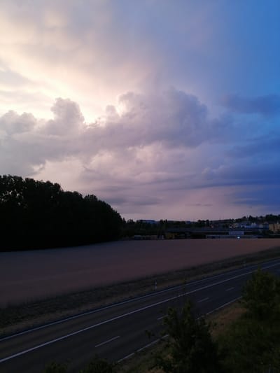 The image captures a twilight scene with a dramatic sky over Ecublens, Switzerland. The sky transitions from soft pinks and purples near the horizon to a deep blue overhead. Heavy, textured clouds dominate the upper portion of the frame. In the foreground, a dark silhouette of a line of trees stretches across the left side of the image. Adjacent to the trees is a wide, empty field, appearing a muted brown or tan color, possibly a harvested field. A dark asphalt road with white lane markings runs diagonally from the bottom center towards the right, separating the field from more vegetation. In the distance, a low-rise industrial or commercial area with several buildings is visible, one of which has signage. The overall atmosphere is serene and somber, characteristic of the end of the day.