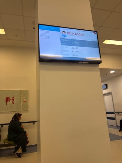 The image shows a digital display screen in a hospital waiting area. The screen displays patient information including appointment numbers, names, and estimated wait times. It indicates that the location is Ankara Bilkent Şehir Hastanesi, which is a city hospital in Ankara, Turkey. The display also shows the date and time as 12.06.2026 / 13:06. Below the main display, there is a smaller screen or label that says "STANESİ". To the left of the display screen, a woman wearing a headscarf and a dark green robe is sitting on a green chair, looking to her left. Behind her, on the wall, is a fire safety sign depicting a coiled fire hose and instructions. The environment appears to be an indoor hallway or waiting room with tiled floors and white walls. A fire alarm detector is visible on the ceiling.